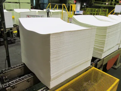 A large stack of blank white paper sheets on an industrial conveyor belt inside a factory setting.