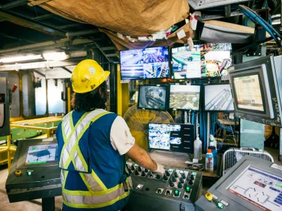 A worker in a yellow hard hat operates control panels and monitors multiple screens in a factory control room.