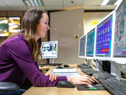A woman in a purple sweater works at a computer station with multiple monitors displaying various data and graphs in a control room setting.