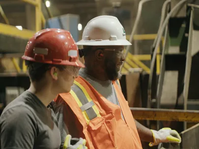 Two workers in a factory wearing hard hats and safety gear stand side by side, observing equipment. One wears a red hard hat, the other a white one with an orange safety vest.
