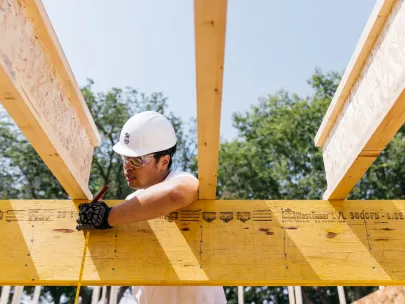 A construction worker wearing a white hard hat and gloves marks measurements on a wood beam using a pencil at a construction site.