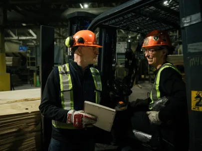 Two workers wearing orange helmets and safety vests converse near a forklift in a warehouse with stacks of plywood in the background.