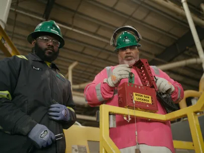 Two workers in safety gear standing indoors; one holds a red lockbox labeled "BROS SAW LOCKBOX.