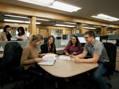 Four people sit at a table reviewing documents, with two people in the background browsing files. Office setting with cubicles and overhead lighting.