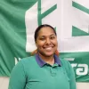 Person smiling in front of a green and white flag, wearing a green shirt and jeans.