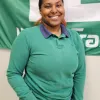 Person smiling in front of a green and white flag, wearing a green shirt and jeans.