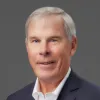 An older man with gray hair, wearing a white shirt and dark blazer, poses against a gray background.