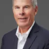 An older man with gray hair, wearing a white shirt and dark blazer, poses against a gray background.