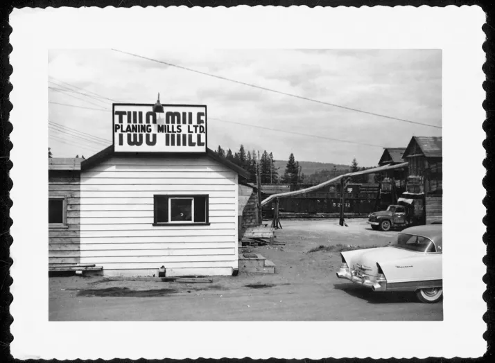 Black and white photograph of a lumber mill with a sign reading "Planing Mills Ltd." A vintage car is parked in front of the building. The background shows additional mill structures and machinery.