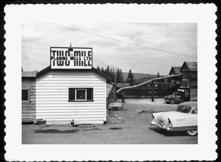 Black and white photograph of a lumber mill with a sign reading "Planing Mills Ltd." A vintage car is parked in front of the building. The background shows additional mill structures and machinery.