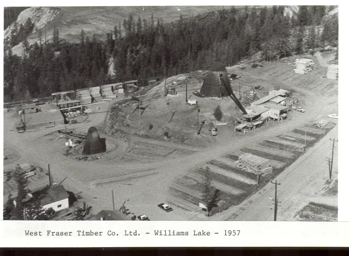 Aerial view of West Fraser Timber Co. Ltd. site in Williams Lake, 1957, showing lumber stacks, industrial structures, and surrounding trees, with vehicles and buildings visible.