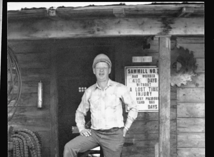 A man stands on the wooden porch of a building, one foot resting on a chainsaw. A sign on the wall behind him displays safety and operational information for "Sawmill No. 1.