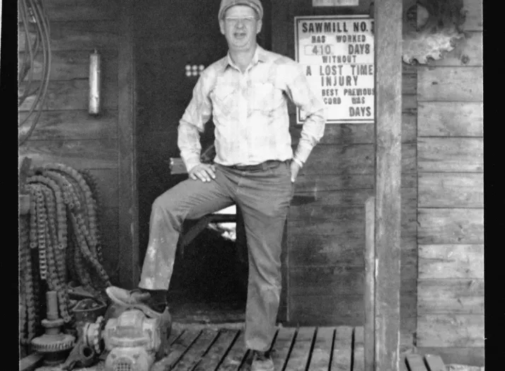 A man stands on the wooden porch of a building, one foot resting on a chainsaw. A sign on the wall behind him displays safety and operational information for "Sawmill No. 1.