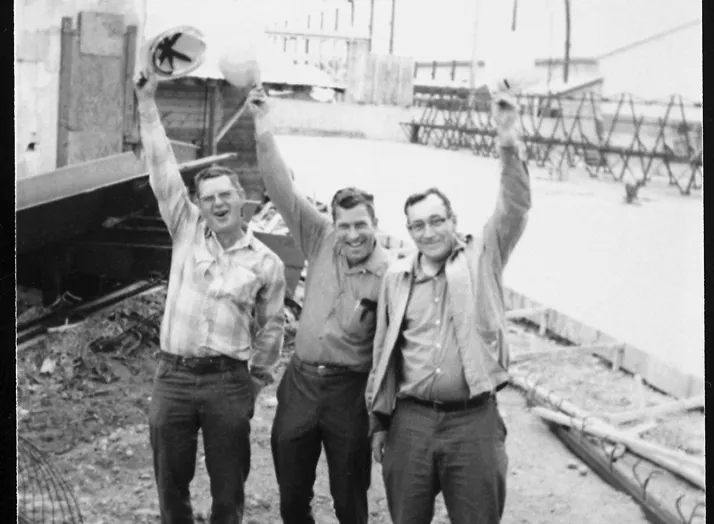 Three men in work attire cheerfully hold their hard hats above their heads at a construction site with scaffolding and building structures in the background.