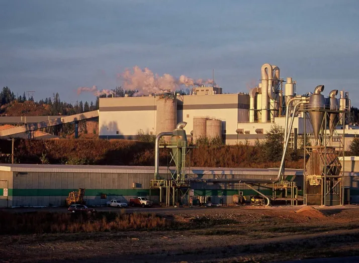 An industrial facility with multiple buildings and large machinery emits smoke from stacks. The surrounding area includes cars and trees, with mountains visible in the background.