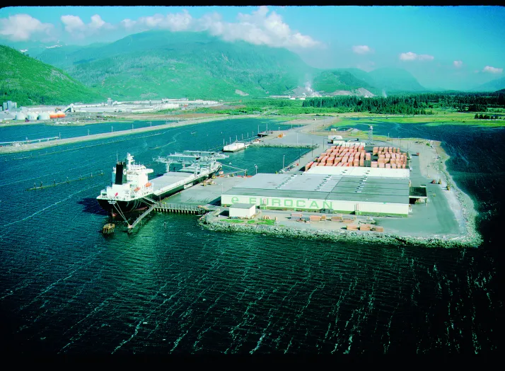 Aerial view of a docked ship at a large waterfront industrial facility labeled "EUROCAN," surrounded by mountains and forests.