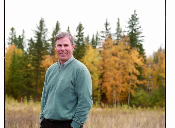 A man stands in an open field with colorful autumn trees in the background, wearing a green sweater and dark pants, hands in pockets, and smiling at the camera.