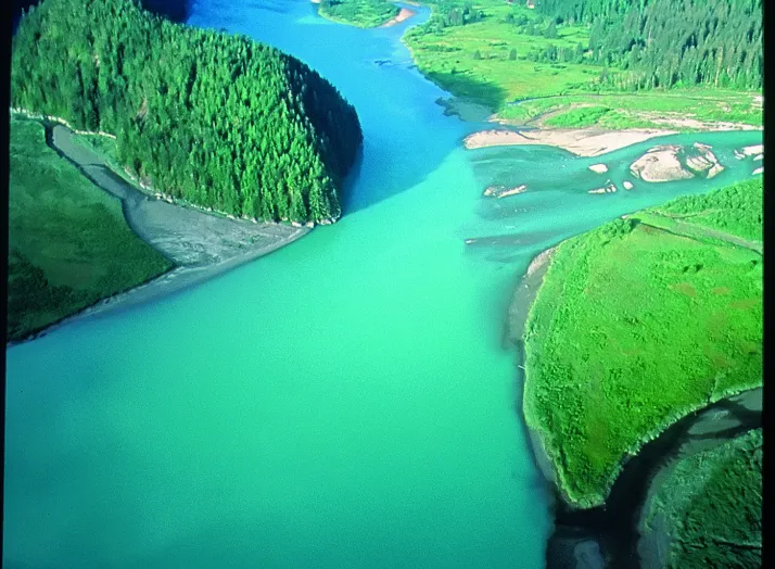 Aerial view of a winding turquoise river flowing through a lush green valley, surrounded by forested mountains.