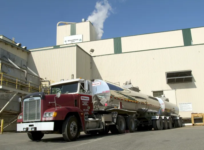 A large tanker truck is parked in front of an industrial building with a tall smokestack emitting white smoke.