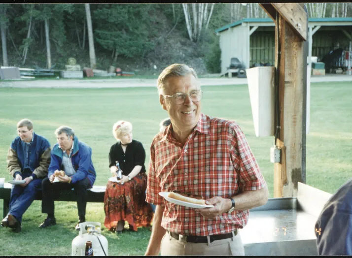 An elderly man in a red plaid shirt holds a plate with food, smiling at an outdoor gathering. Four people sit on a bench in the background, with trees and a building visible.