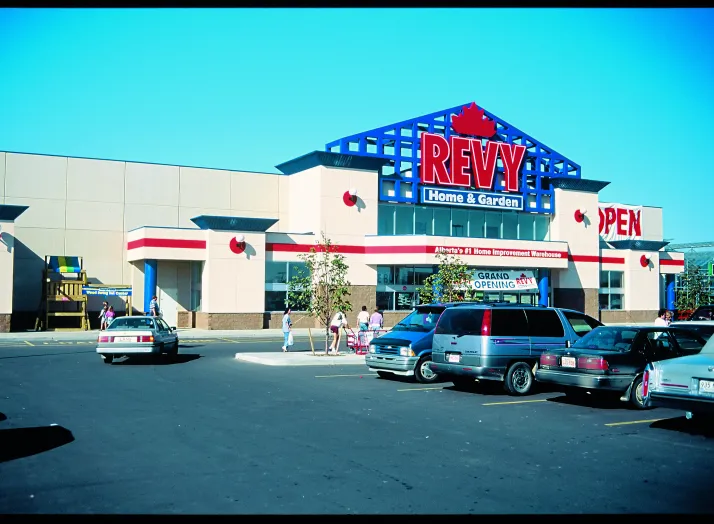 A Revy Home & Garden store exterior with cars parked in the lot and several people walking towards the entrance on a sunny day.