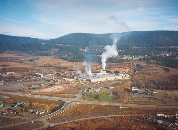 Aerial view of a large industrial plant with smoke rising from chimneys, surrounded by a vast landscape of hills and roads.