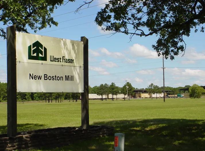 Sign for "West Fraser New Boston Mill" stands in a grassy field with trees and industrial buildings in the background.