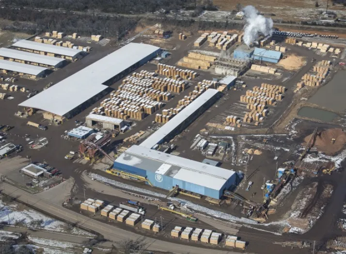 Aerial view of a large industrial lumber facility with multiple buildings, stacks of wooden logs, and visible machinery. Smoke is emitting from one section of the plant. Surrounding area includes roads and vegetation.
