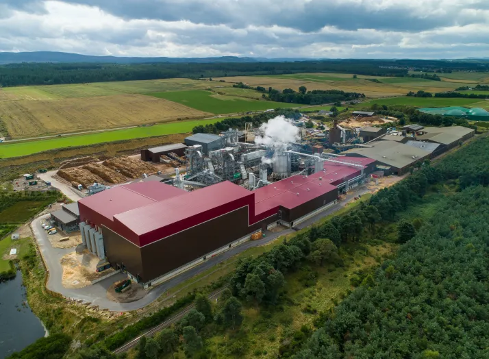 Aerial view of an industrial plant with red-roofed buildings surrounded by fields and forests under a cloudy sky.