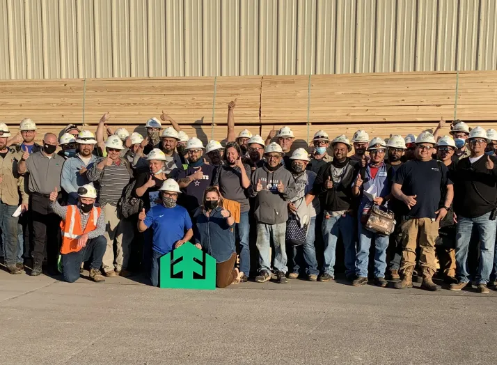 Group of construction workers in hard hats and safety gear standing together in front of stacked lumber, posing for a group photo.