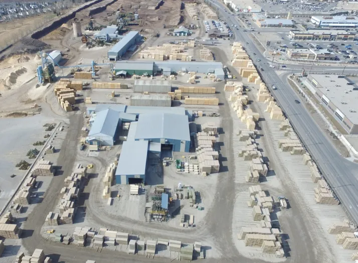 Aerial view of a lumber yard featuring stacks of wooden planks, multiple buildings, and nearby roads and industrial areas in the background.